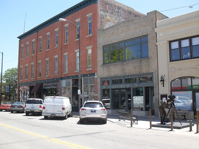 Multi-storied historic storefronts in downtown Loveland with diagonal cards parked. On the ride side of the image a statue of a person with a film camera sits on the sidewalk near a bench.