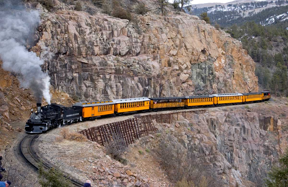 The black and yellow cars of the Durango & Silverton Narrow Gauge Railroad hug the rocky ledge of a mountain. 