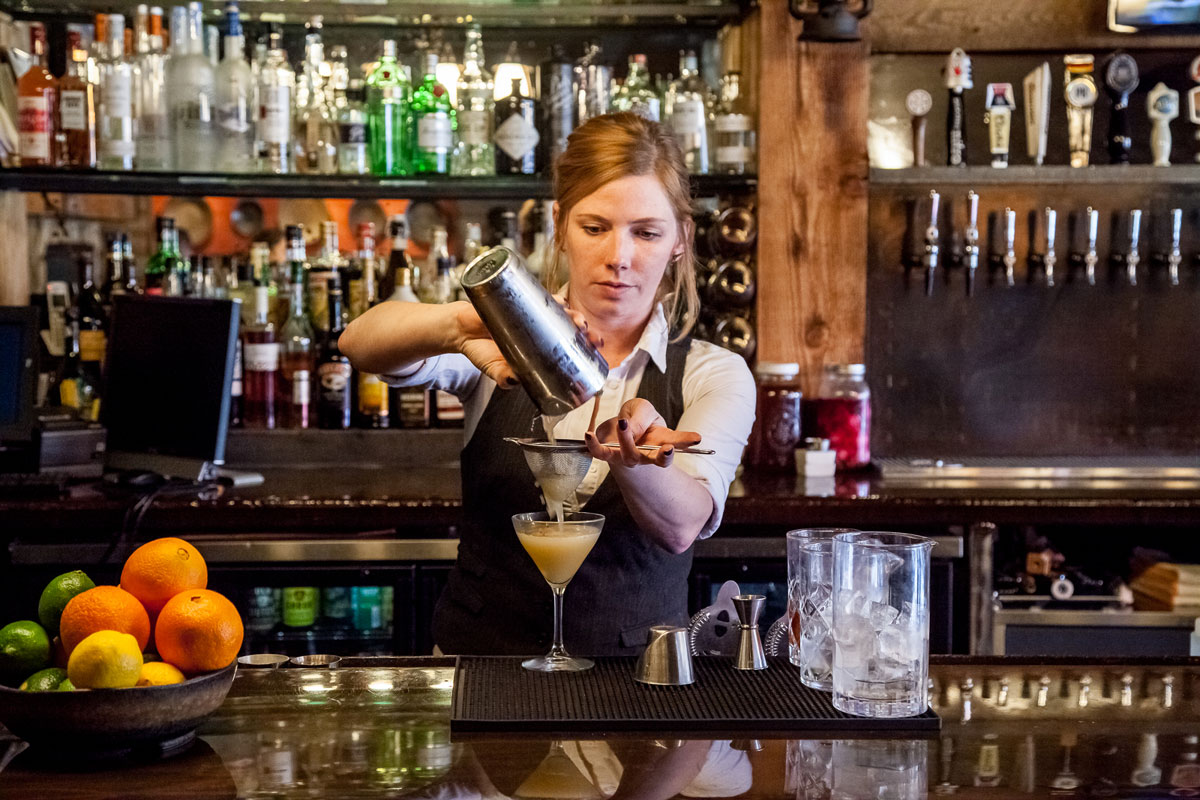 A bartender pours a creamy drink from a metal shaker through a strainer into a martini glass. She's wearing a vest and there's a bowl of citrus fruit on the left. Behind her are wooden shelves filled with alcohol bottles at Miners Saloon.
