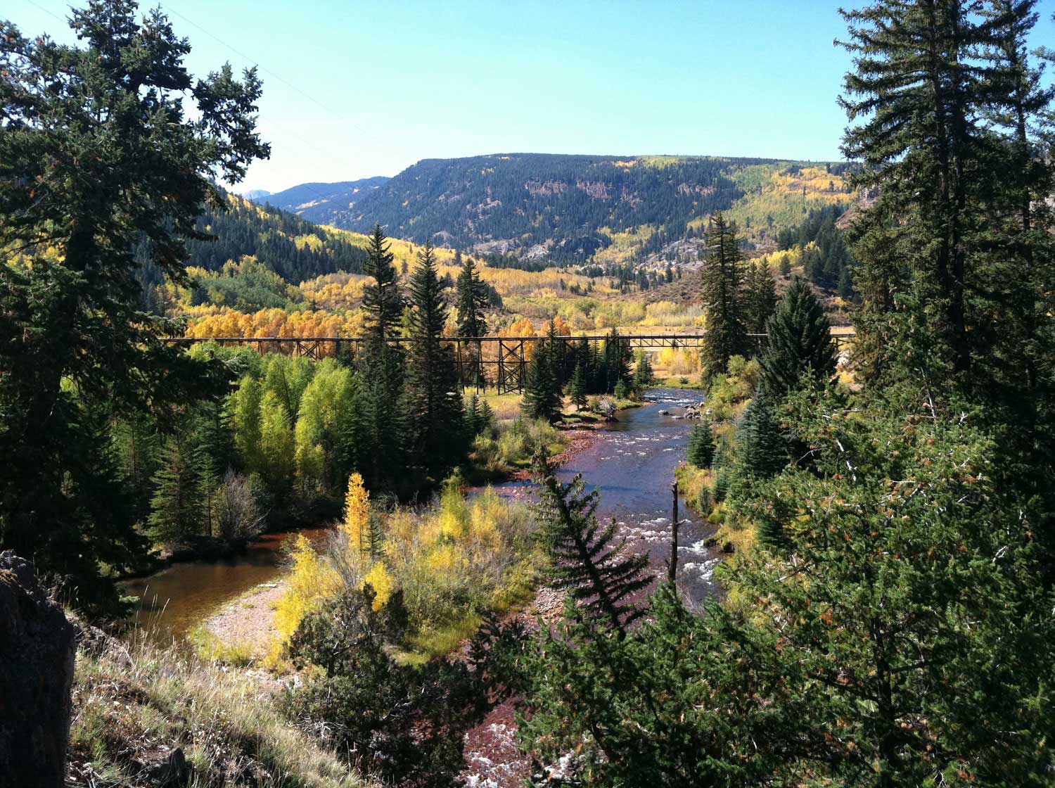 A railroad bridge sits among evergreen and golden-hued trees above a river with mountains in the distance.