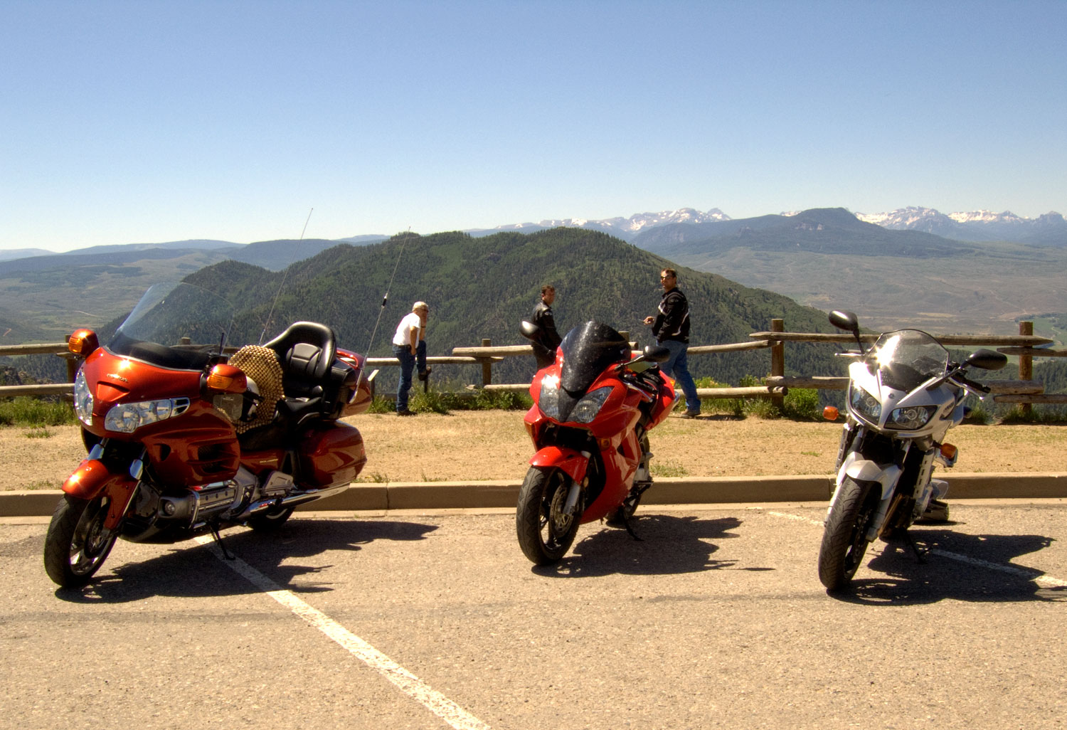 Three motorcycles stand empty in a parking lot overlooking Colorado's San Juan Mountains. Three riders send by the wood-pole fence and admire the scenic view.