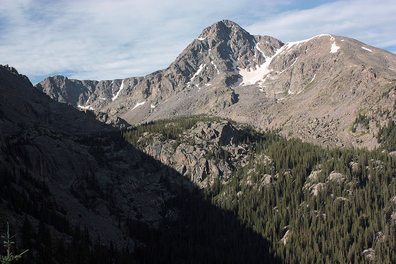 The rocky peak of Mount of the Holy Cross sits with snow nestled in a nook. Beneath it there are rocky outcroppings with evergreen trees.