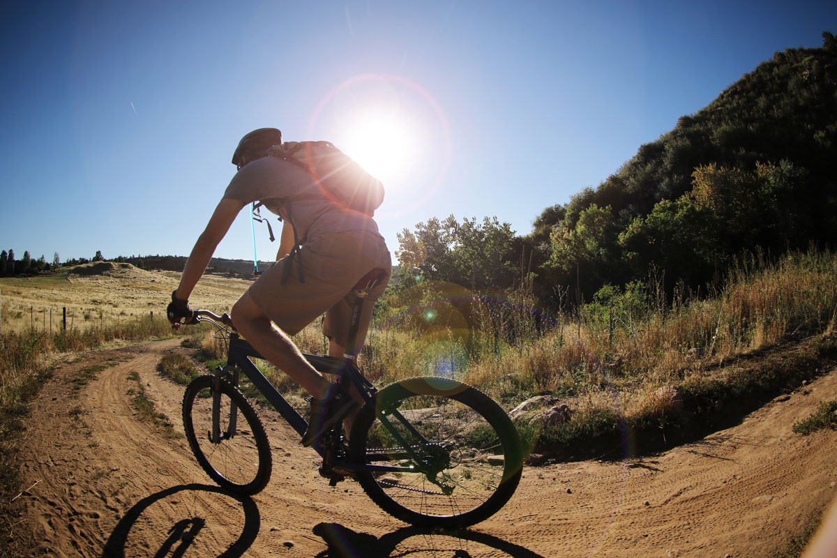 A person on a mountain bike thunders along a dirt path on a blue-sky day with the glowing sun looking down. On the right there are green trees.