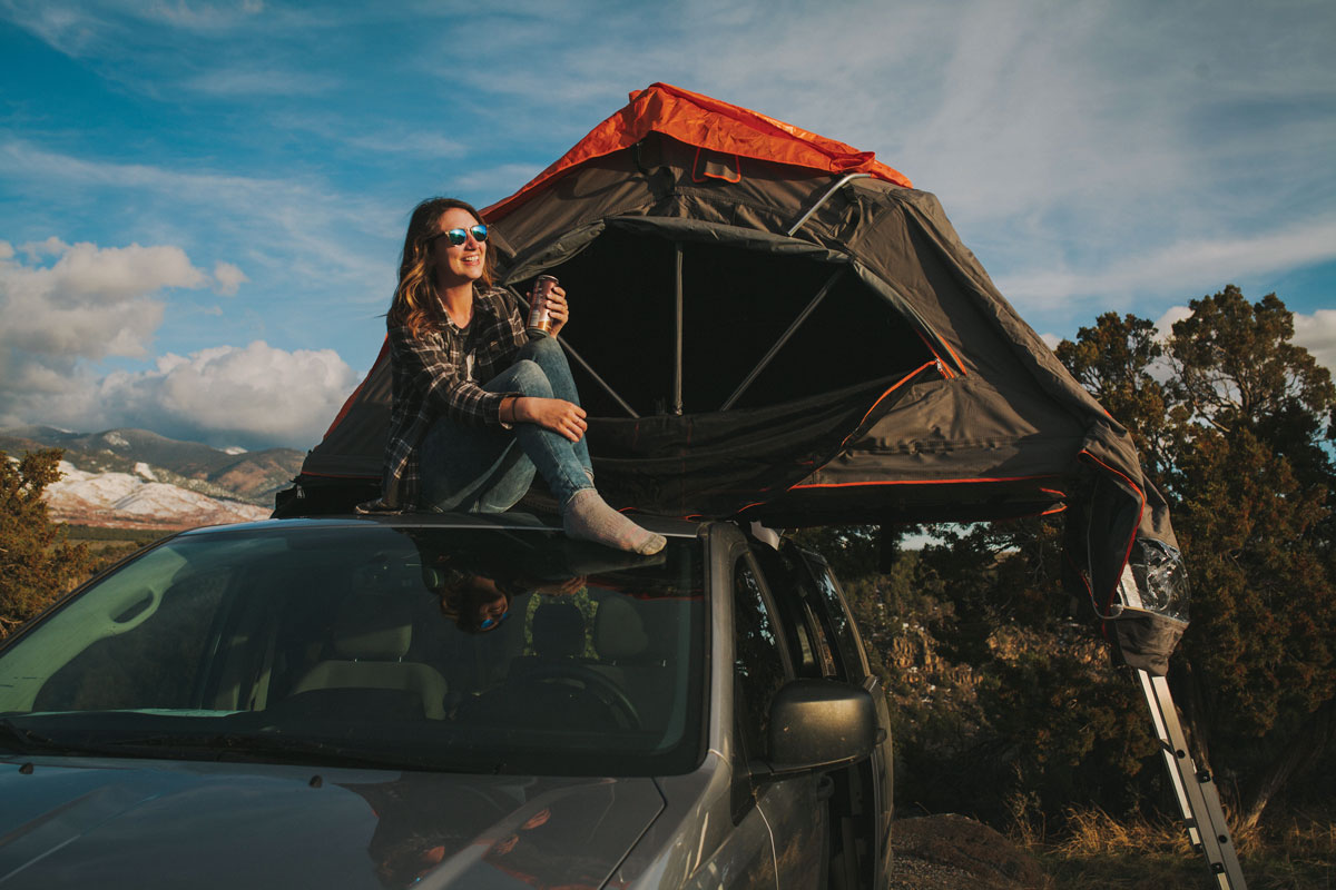 A woman sits atop a Native Campervan — a van with a fold-out tent that rests partially on the van, with the other side on a platform with a ladder down to the ground