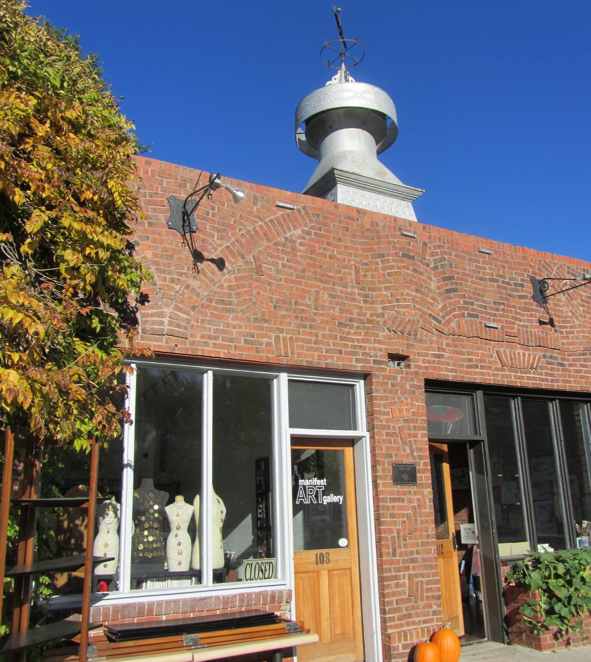 A red-brick building with a door that says "Manifest Art Gallery" sits under a blue sky.