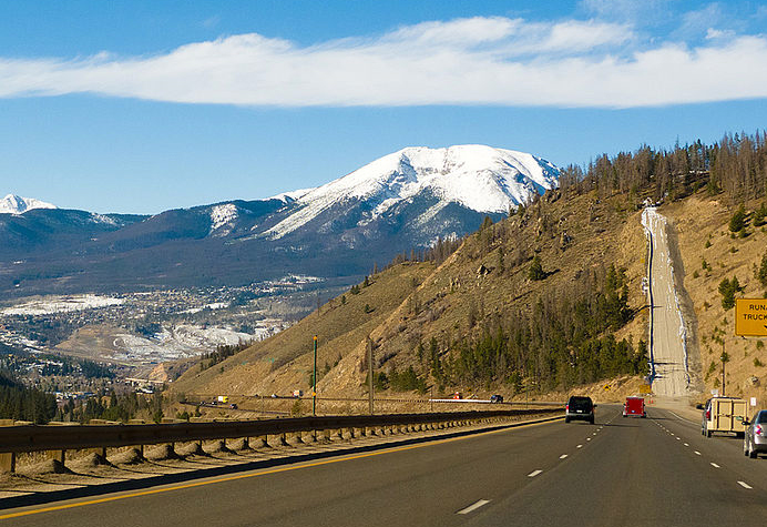 A three-lane road with cars on it winds down into town. There are snowcapped mountains in the distance with blue skies and white clouds. A runaway truck ramp sits to the right on a brown hill with evergreen trees.
