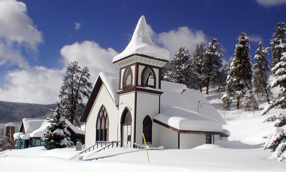 Bright-white snow blankets the historical Pitkin Community Church in downtown. There's a blue sky with white clouds and evergreen trees.