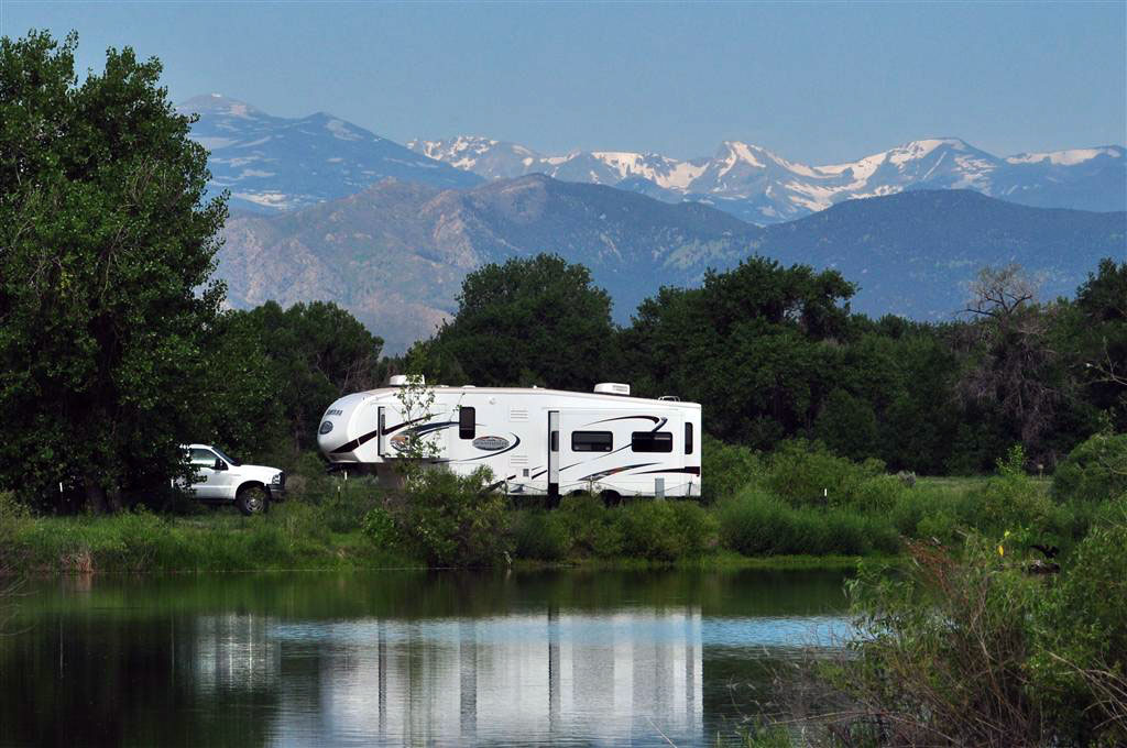 A white RV sits in the middle of a green grass and trees. It's sitting in front of a lake that's reflecting its image. In the background there is a snow-capped mountain range.