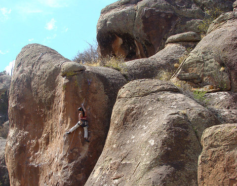 A solo climber has climbed halfway up a reddish rock in a rocky group that has little tufts of green grass.