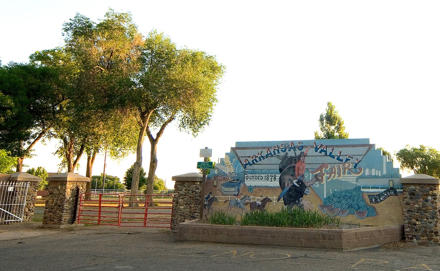 The sign of the Arkansas Valley Fair is painted with a bull bucking its rider. There's a red metal gate between two stone entrance posts and the golden sunlight is upon green-leafed trees.