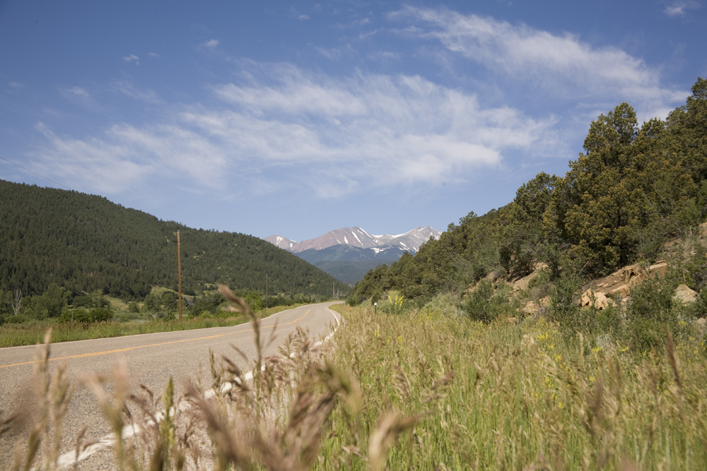 On a summer's day the road along the Highway of Legends Scenic and History Byway stretches out with evergreen trees on both sides and tall grasses. In the distance snow-capped mountains rise up.