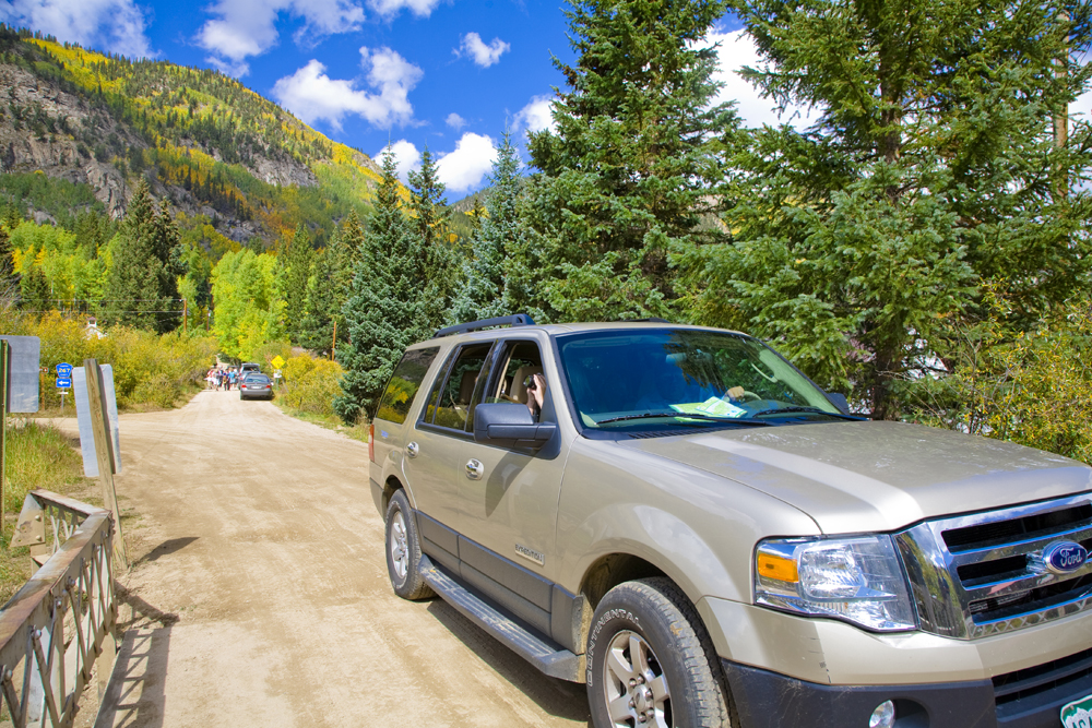 A car drives across a small bridge with mountains and blue sky in the background