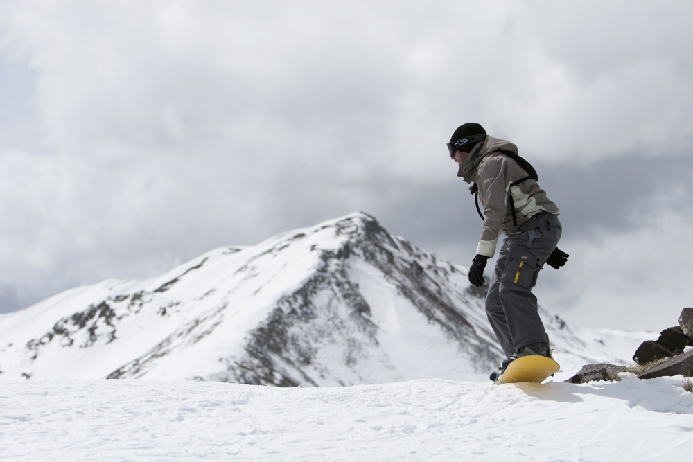A grey-clothes-wearing snowboarder boards on a yellow snowboard with a snow-covered mountain peak in the background. 
