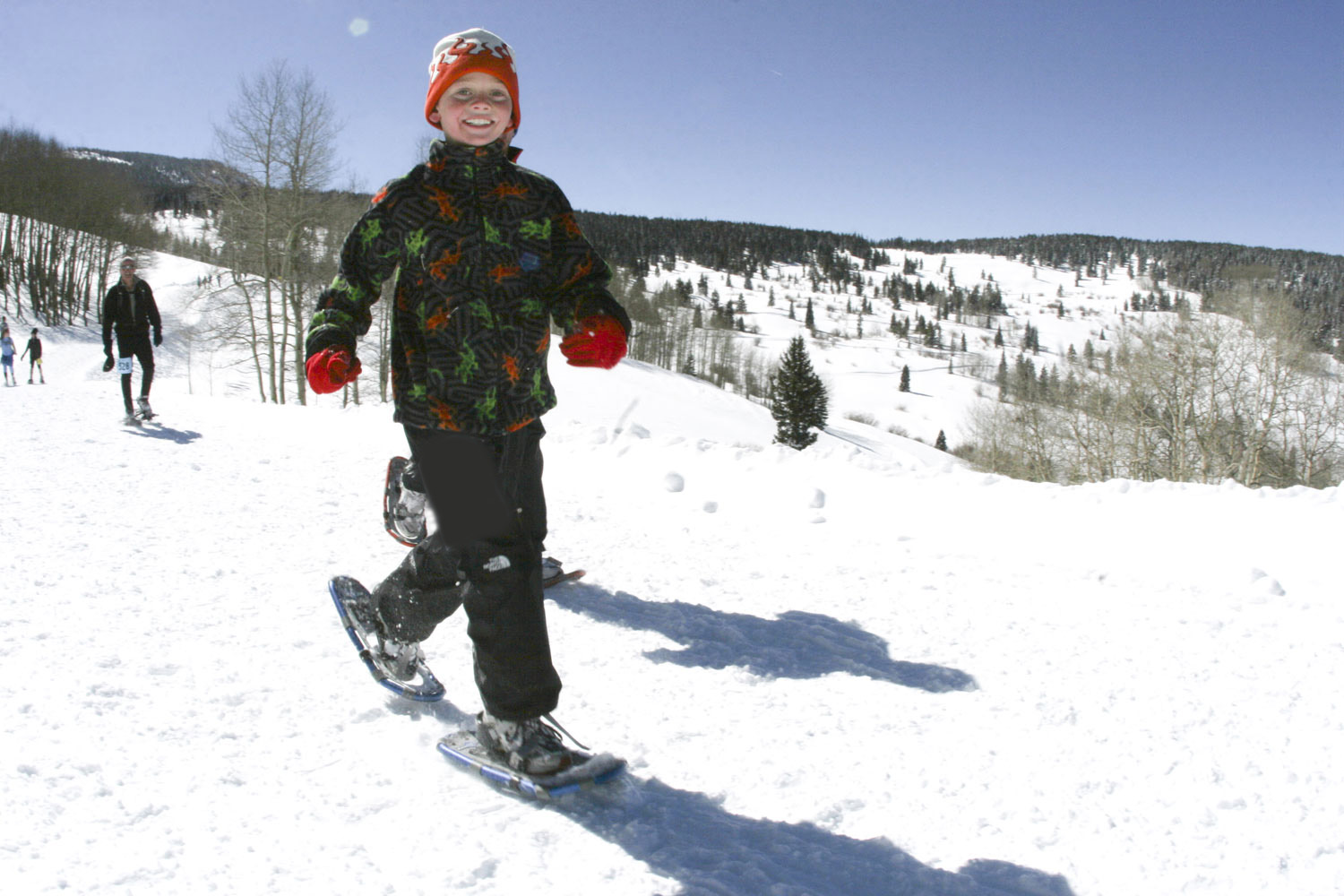 Child snowshoeing on a blue sky winter day. Small trees with no leaves dot the background and a four other people are snowshoeing in the background.