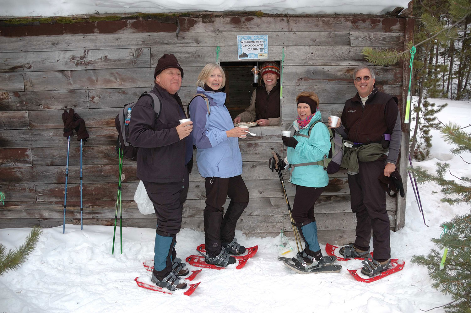 Four people pose with the hot chocolate stand during a snowshoe adventure in State Forest State Park near Walden. The stand is grey wood and the worker is standing in the square window also smiling.