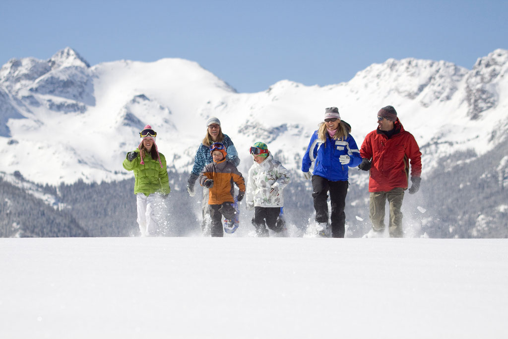 Four adults and two children in colorful ski jackets snowshoe over a snowy hill with soaring snow-covered mountains in the background. 