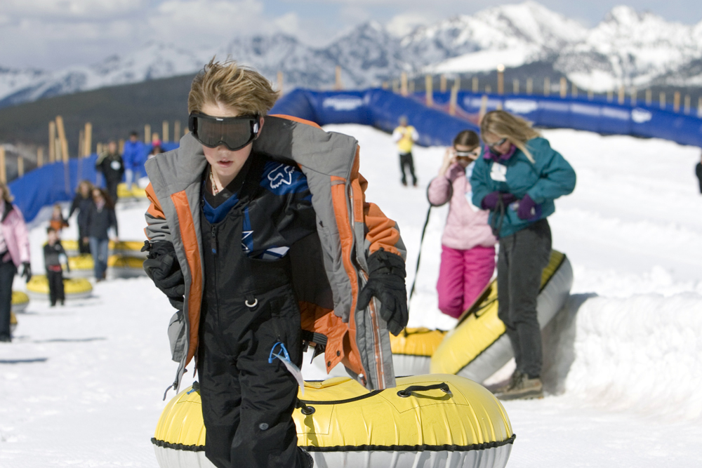 A boy tows his yellow tube on a rope up a hill to get ready to head down it again; several other riders are lined up behind him