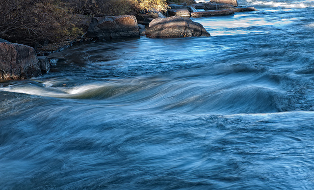 The flowing South Platte River moves. The bank to the right has boulders and scrubby green bushes.
