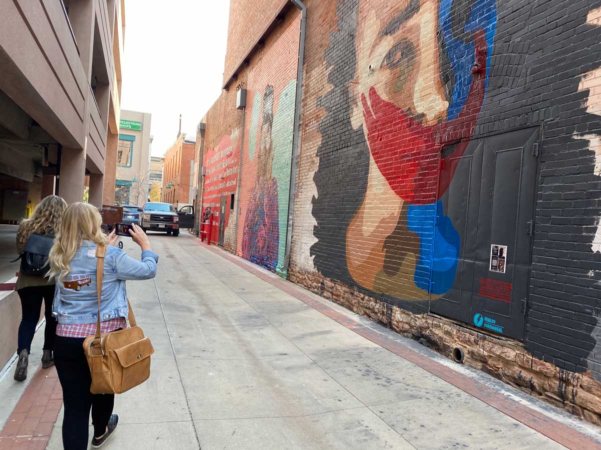 A woman takes a photo of StreetWise, a Mural by Gregg Deal which depicts a woman with bright red and blue colors covering her mouth