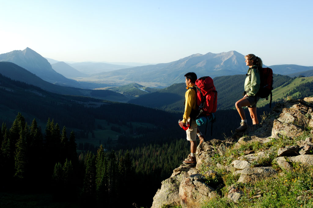Two people wearing big hiking backpacks stand at a cliff's edge and look across a canyon to blue peaks shaded by fog.