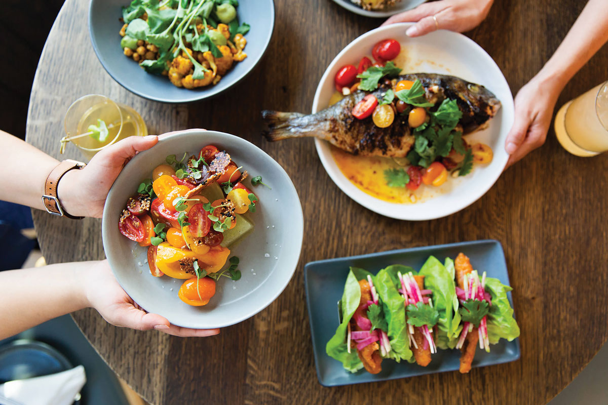 A person passes a gray bowl of salad topped with watercress over a dark wood table at The Kitchen in Boulder, Colorado. Other dishes include a cooked whole fish, lettuce tacos and curry cauliflower.
