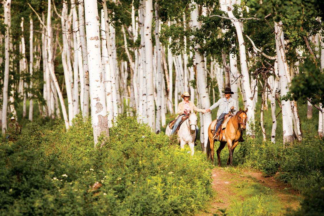 A smiling couple holds hands as they ride horses down a trail in an aspen forest at The Home Ranch in Clark.
