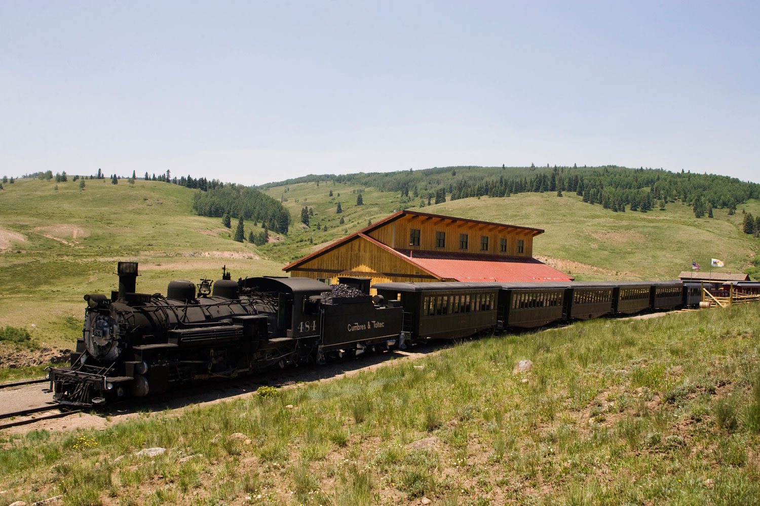 On a summer day with green hills, in the middle the historic Cumbres & Toltec Scenic Railroad train is pulling out of the depot.