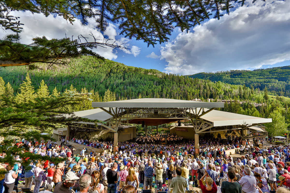 A crowd lined up in front of an amphitheater with evergreen forest behind it