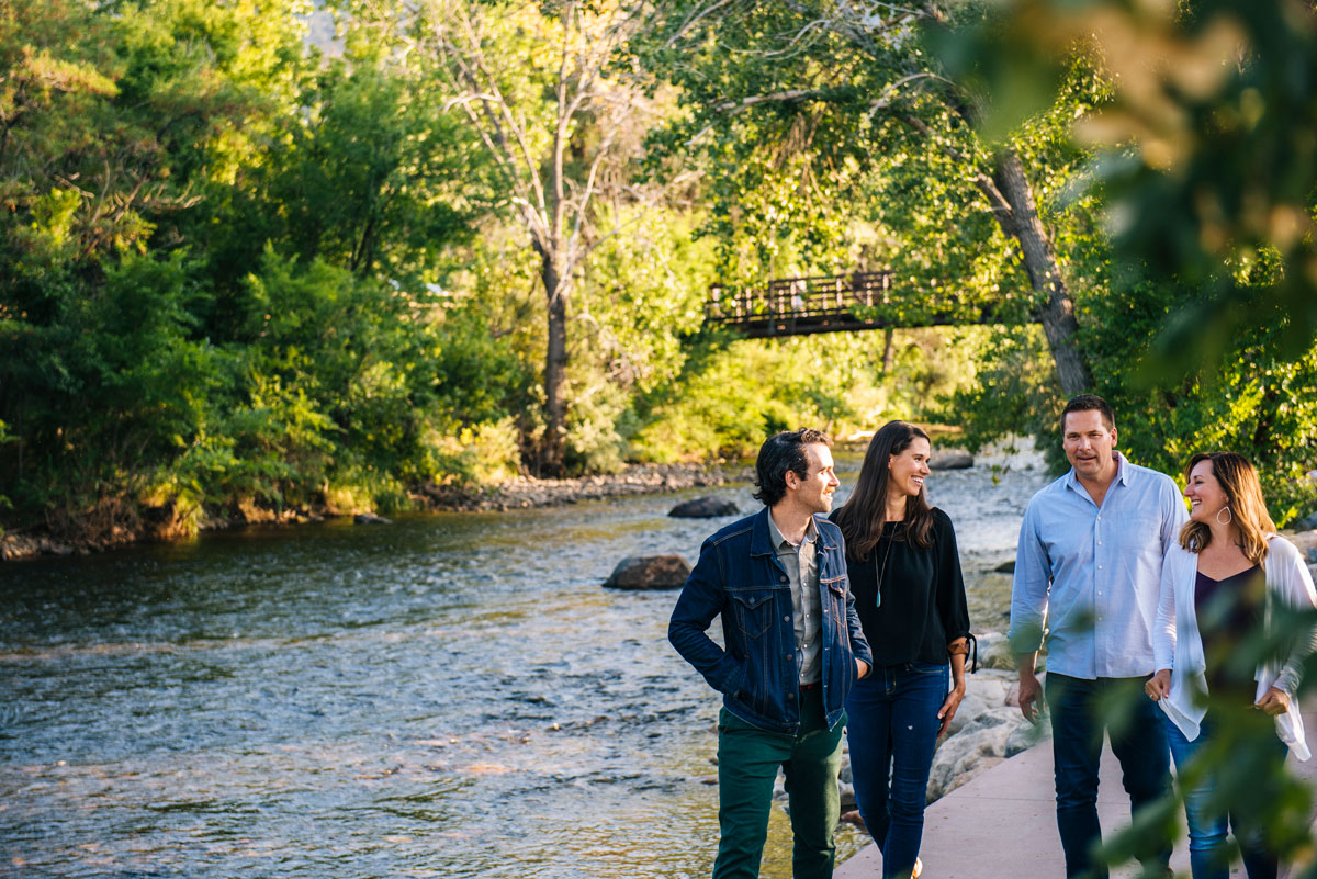 Four people stroll along Clear Creek in the summer. The river is surrounded by verdant trees and bushes. In the background there's a pedestrian bridge over the water. 