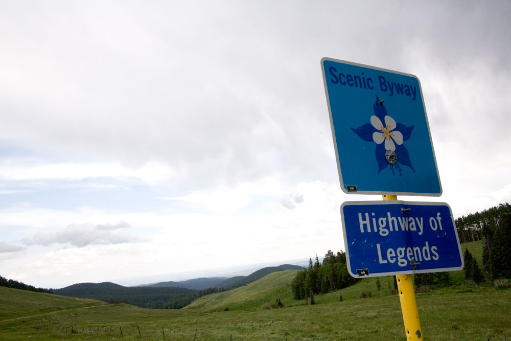 A green, mountain vista along the Highway of Legends Scenic and Historic Byway in southeast Colorado. On the right a metal square sign with a flower on it says "Scenic Byway." Below that on the yellow sign pole is a blue rectangular sign that says "Highway of Legends."