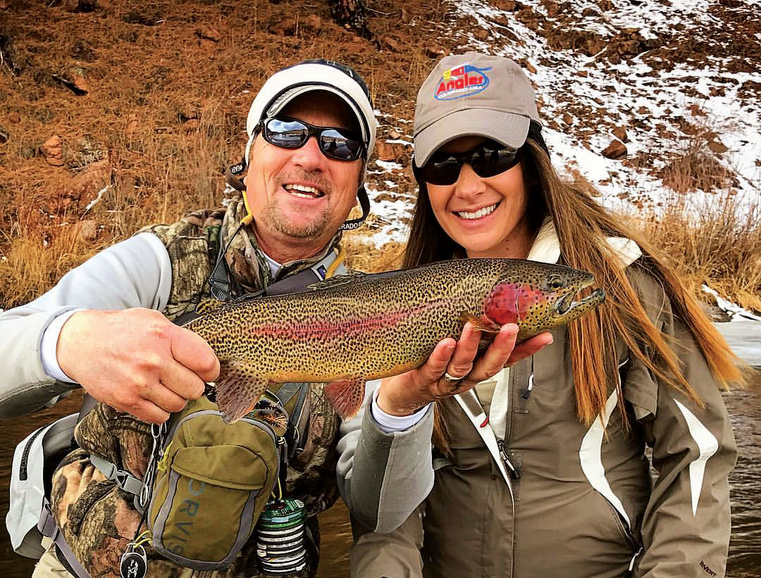 Two wintertime anglers smile and pose with a recently caught freshwater on a river in Colorado. The fish is yellow with brown speckles a red stripe down its side and a blush spot around its gills.