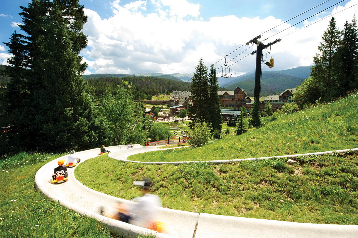 People sitting on yellow mats fly down a white half-tube slide that curves down a steep mountainside at Winter Park Resort in Colorado.