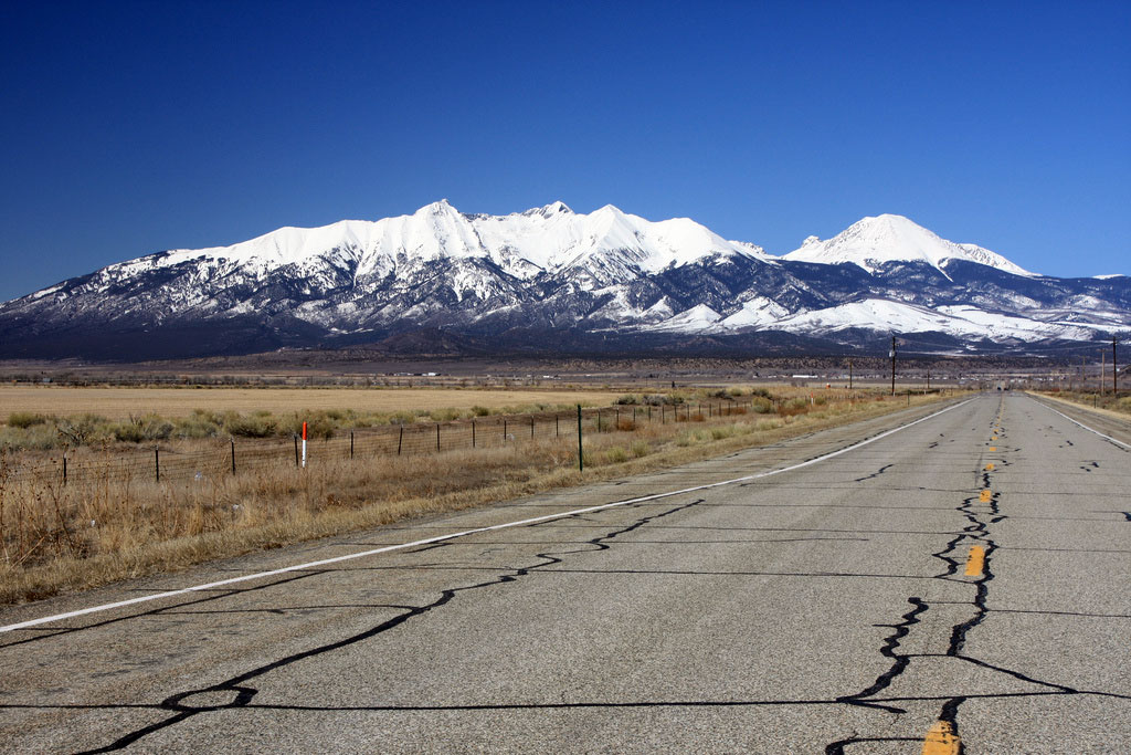 A massive mountain range with snow-covered peaks sits under a cloudless, bright-blue sky with a cracked road and brown grasses leading up to it. 