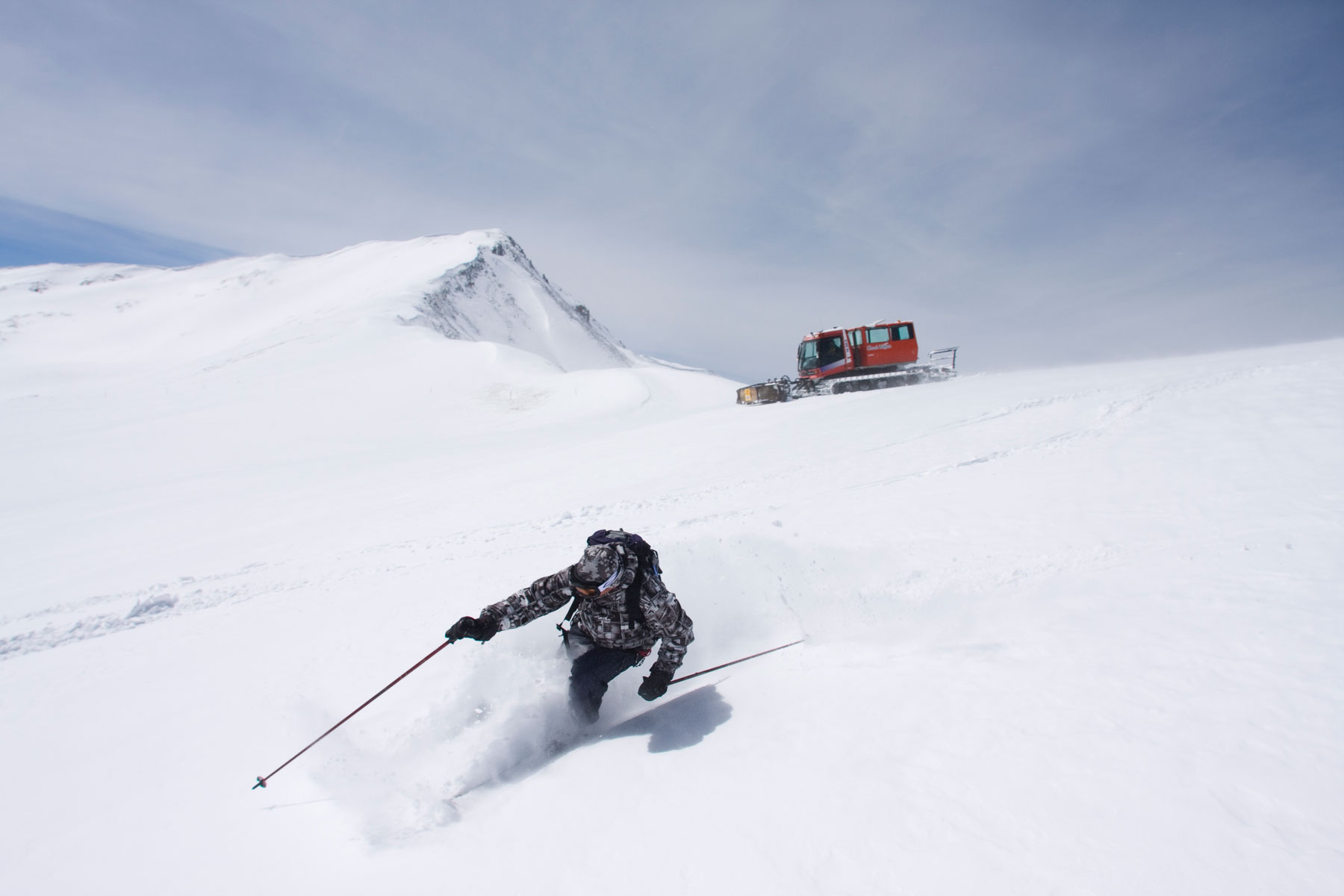 A skier makes their way down a snowy slope with a red snowcat sitting atop the ridge in the distance.