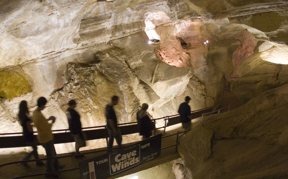 Six people cross a bridge that has a sign that reads "Tour Cave of the Winds" in a rock cave with high ceilings.