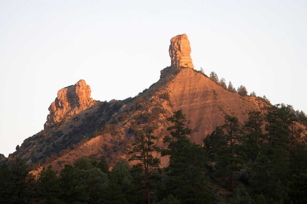 Up against a hazy white sky the sun is setting, lighting up the pillars of Chimney Rock Archeological Area. There are evergreen trees in the foreground.