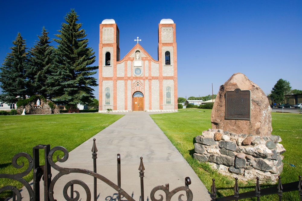 On a summer's day, Our Lady of Guadalupe church sits under a blue sky with green grass and evergreen trees in front. There are two tower's on the church that is outlined in pink brick. On the right is a historical marker made out of stone.