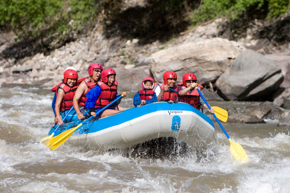 A family rafts in the Colorado River’s whitewater rapids. They are wearing red helmets and red life jackets and are seated in a blue and white inflatable boat. They’re holding paddles with blue poles and yellow paddles. They’re surrounded by big grey boulders and green grass on the banks.