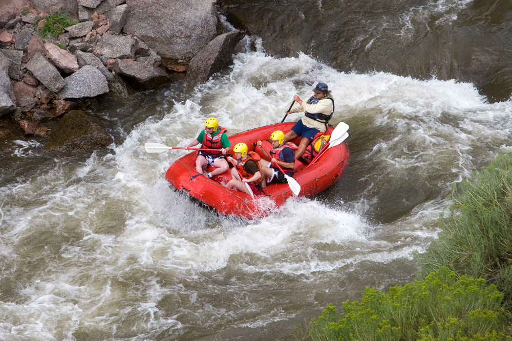 A group of four people whitewater rafts in a red raft down the Arkansas River. There are rocks on the left and a green tree on the right.