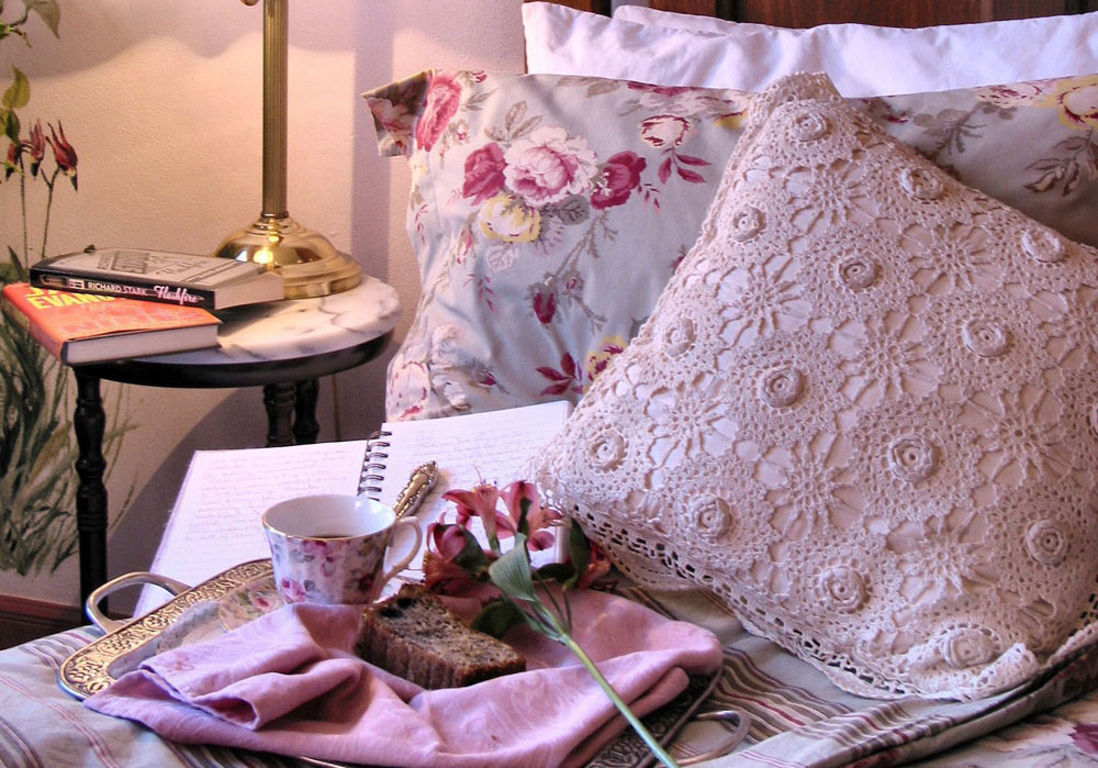 A breakfast tray with a slice of fruit cake, a flowered tea cup and pink flower sits by an open journal with elegant cursive writing on a pillow-ladened bed in Denver, Colorado.
