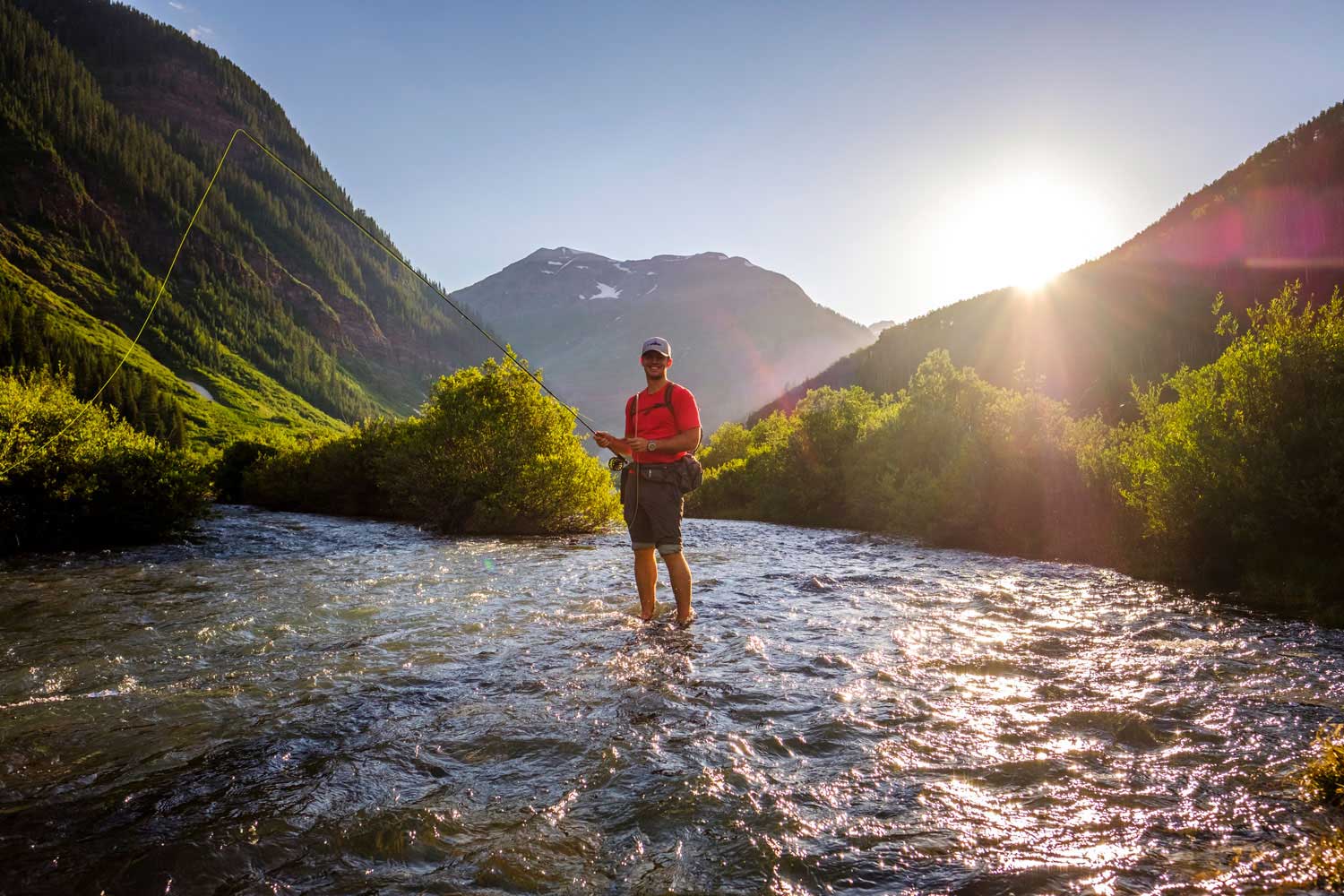 Someone is ankle-deep in a Durango river, casting their fly-fishing line in the waters. Along both of their sides are two tall grassy hills that reach past the top of the photo. The sun peaks over the right side of the hill, and there is a tall, jagged mountain peak behind the person in the photo.