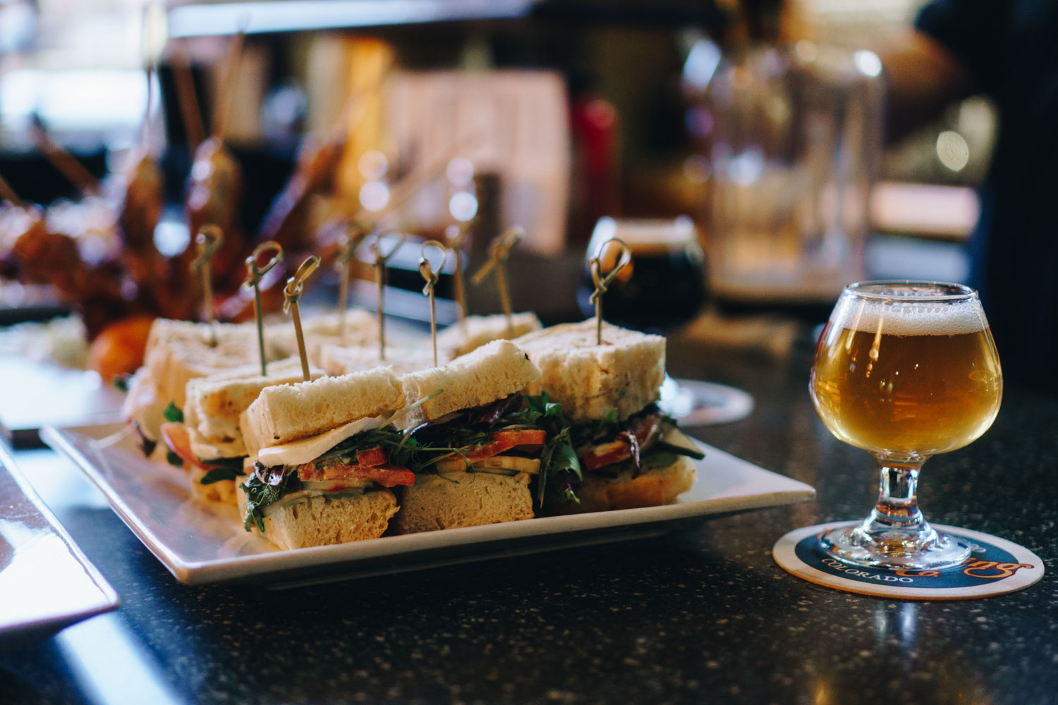 A plate of nine finger sandwiches consisting of white bread, white cheese, greens and a tomato, stuck through with a toothpick. There are two glasses of foaming beer next to the plate.