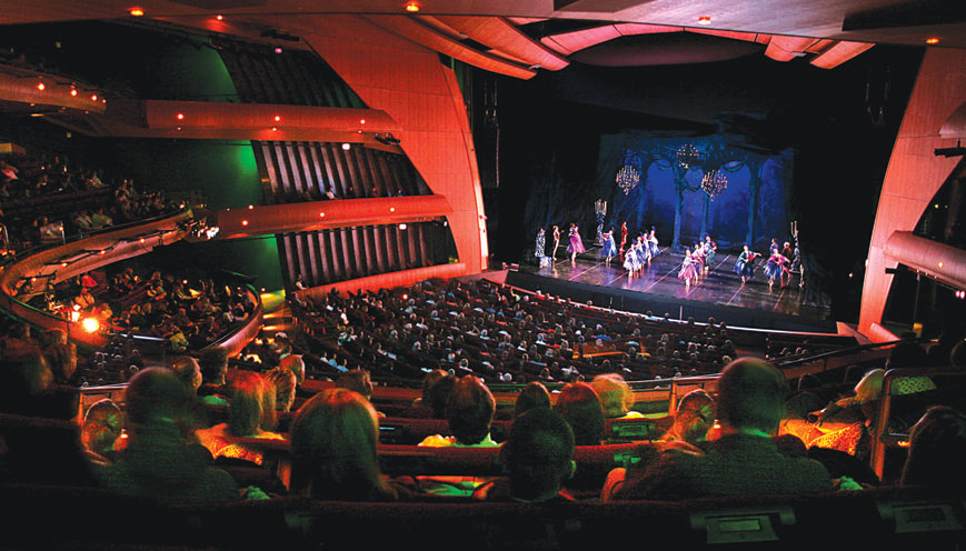Audience seating is lit with red and green lighting at Ellie Caulkins Opera House where dancers perform on a large stage.