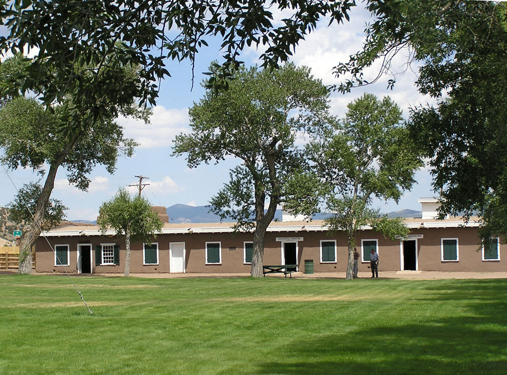 A summer's day at Fort Garland Museum. The one-story fort is painted brown and has a green lawn and there are green-leafed trees. Two people stand near one of the walls.