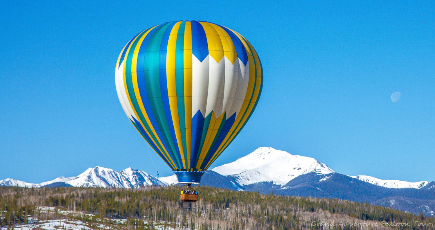 A faint waxing moon and snow-blanketed mountains stand out against a cloudless, bluebird sky in Winter Park. Before them floats a blue, yellow, white and teal hot hair balloon carrying people in a woven basket.