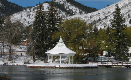 The white historic gazebo in Green Mountain Falls sits on a lake. The ground is covered in snow and the evergreen trees are dusted with snow. Behind them there are snow-covered mountains.