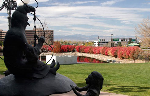 On the left a sculpture of a man sitting above a wondrous child. In the background a green-grass field sits next to a lake with pink flower bushes and in the background there's a hazy view of the Front Range. 