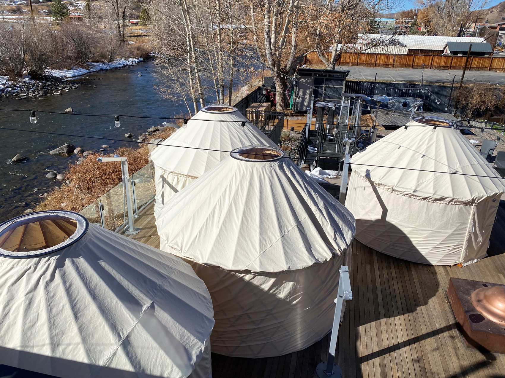 Four off-white yurts with holes at the top of the tents are set up at an event space alongside the flowing body of water in Steamboat Springs in winter.