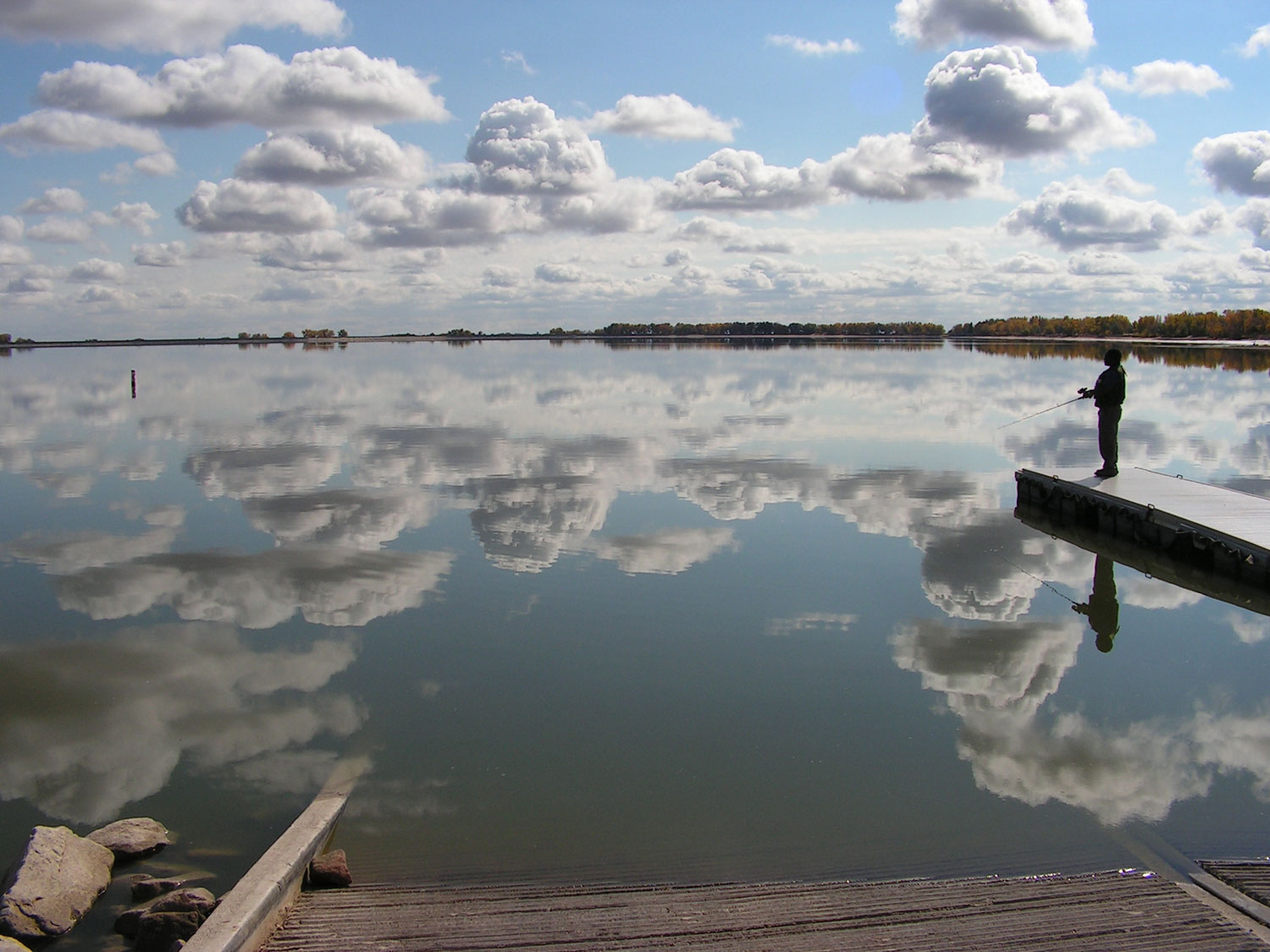A blue sky dotted with puffs of clouds is reflected in the mirror-smooth surface of Jackson Lake near Fort Morgan, Colorado. To the side is the silhouette of an angler on a dock. 