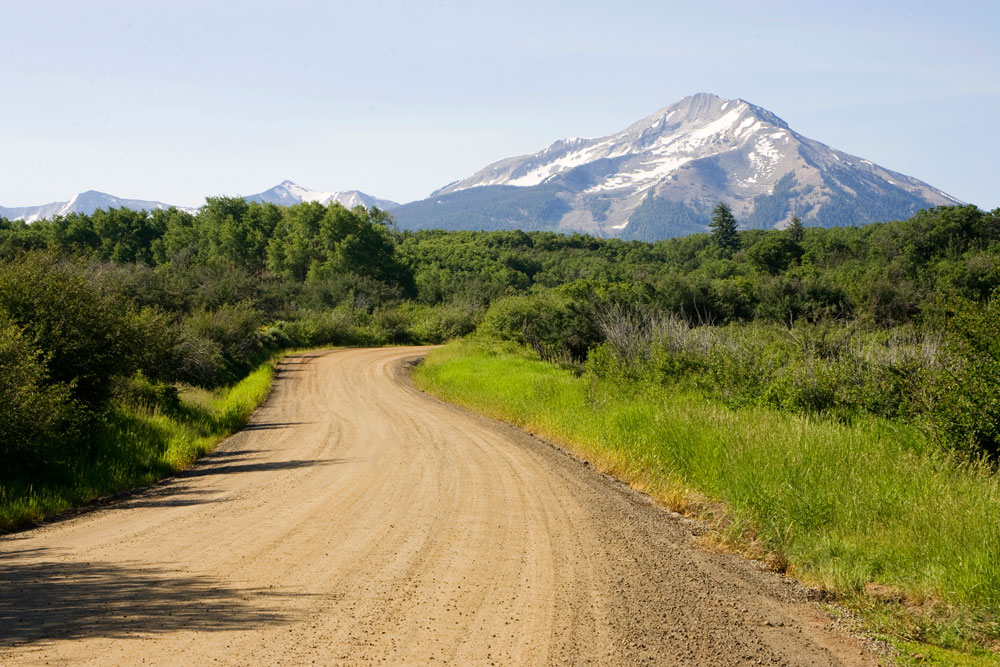 A dirt road is surrounded by green grass and green bushes on a hazy day with snow-covered stone mountains on a route between Somerset and Crested Butte.